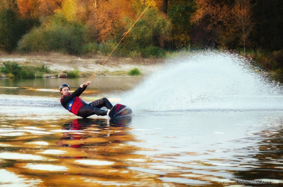 A man wakeboarding on a calm river during autumn, leaning back and carving through the water, creating a large spray against a background of golden trees.