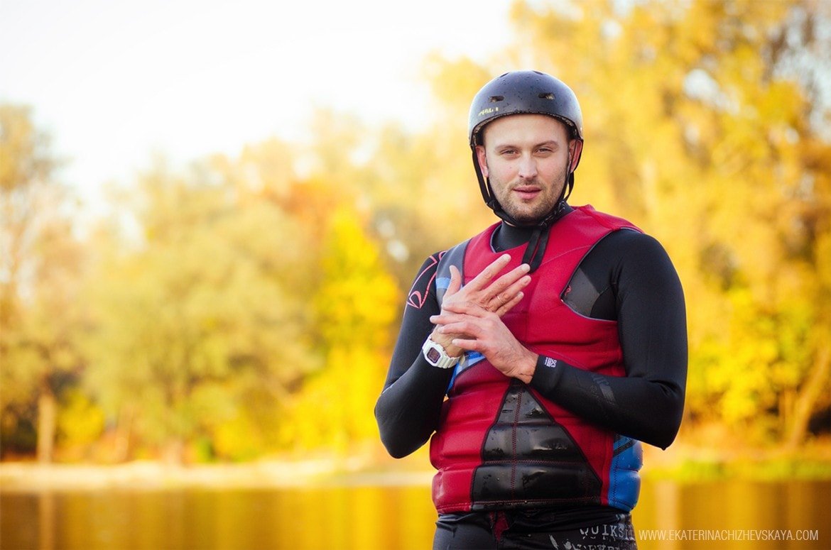 A close-up portrait of a male wakeboarder wearing a black helmet, red life jacket, and a white watch, looking at the camera against a soft-focus autumn landscape.