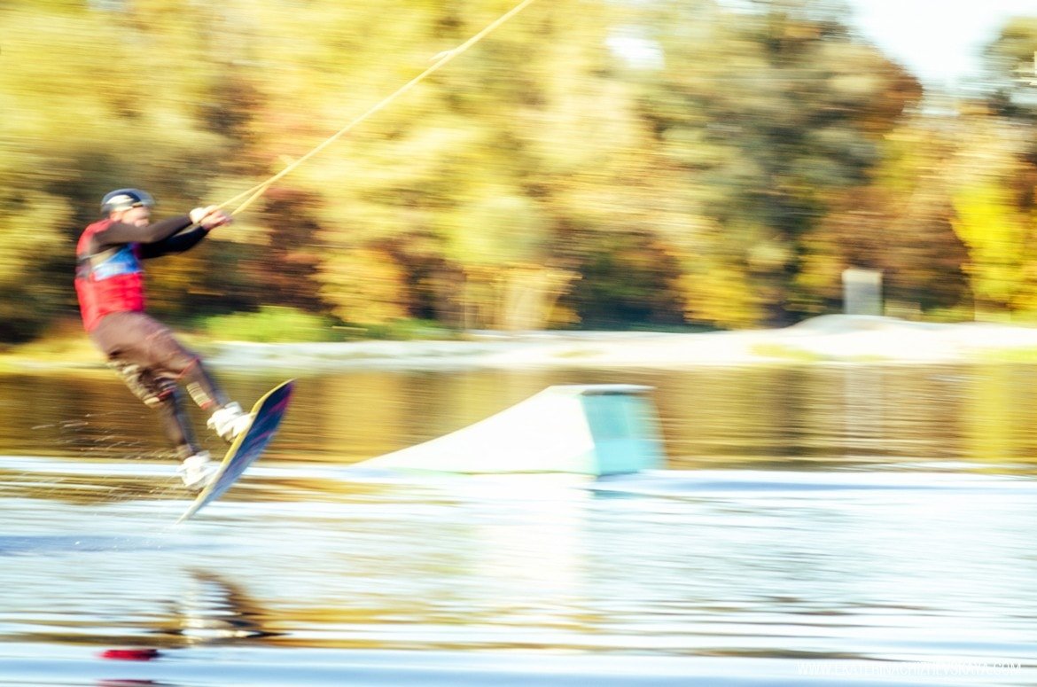 A motion blur action shot of a wakeboarder launching into the air over the water near a ramp, with a golden fall forest blurred in the background.