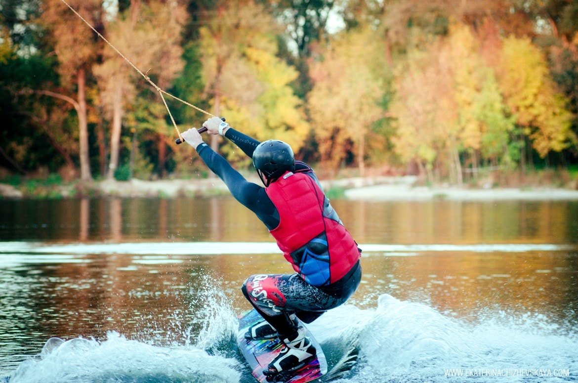 Rear view of a man wearing a red life vest and helmet wakeboarding on a lake, holding the tow rope with autumn foliage in the background.