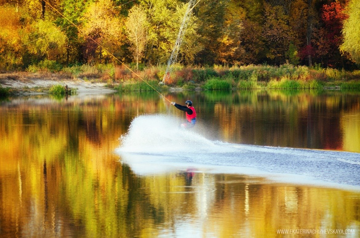 Wide shot of a wakeboarder gliding across glassy water reflecting vibrant yellow and orange autumn trees on the shoreline.