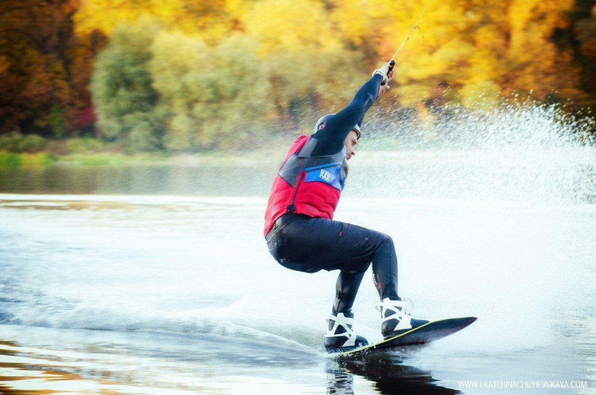 Side view of a wakeboarder in a wetsuit and red vest cutting through the water, creating a big white spray with colorful fall forest blurred in the background.