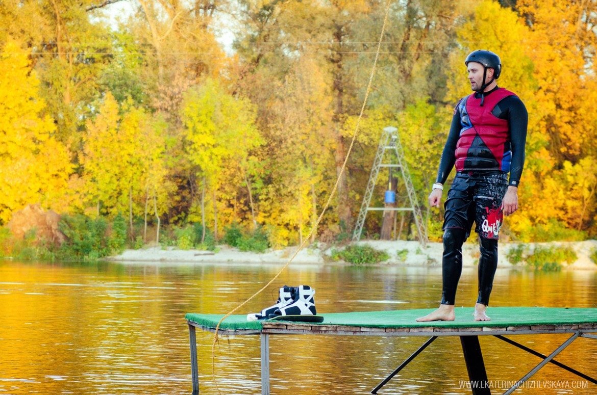 A man in a wetsuit, red life jacket, and helmet standing on a wooden dock next to his wakeboard, preparing to ride on an autumn day.