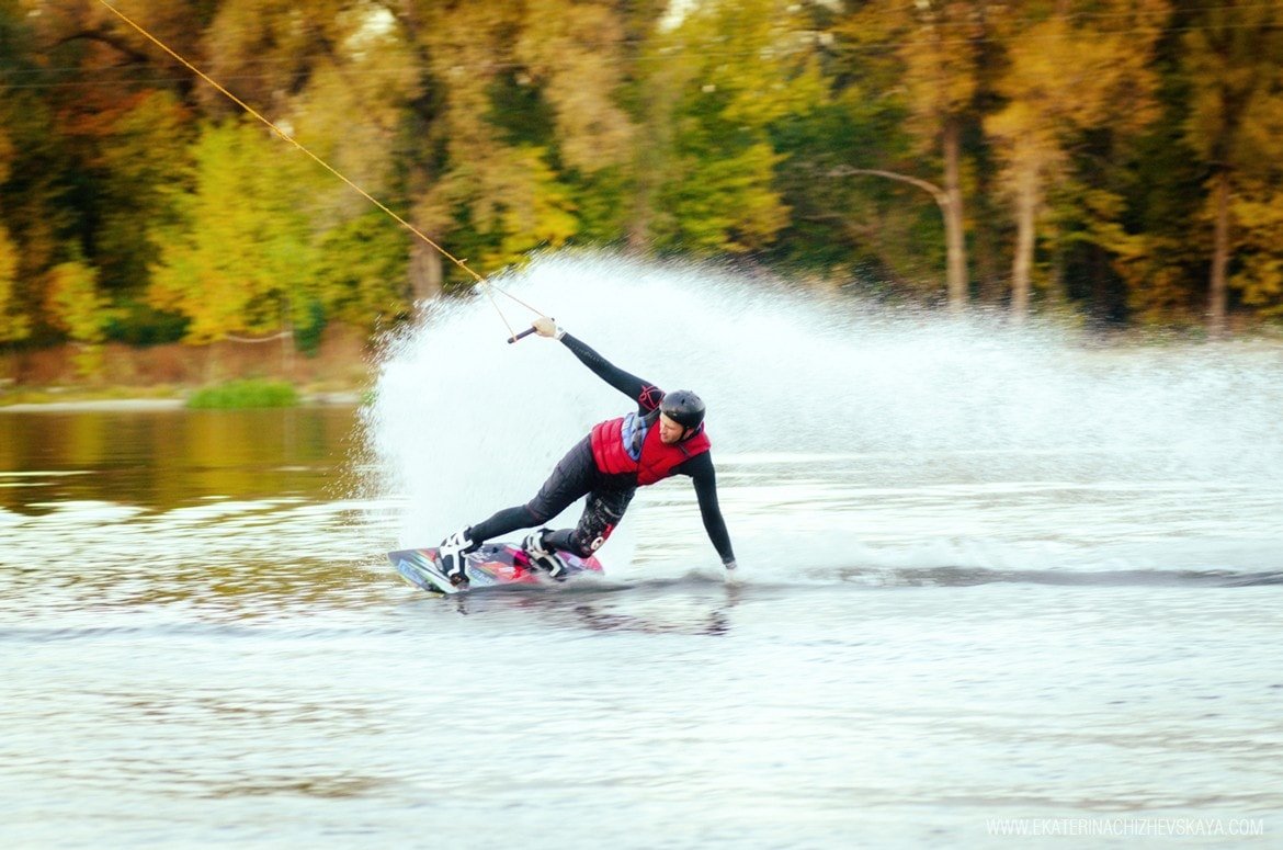 An athletic man in a wetsuit leaning hard while carving on a wakeboard, pulling on the tow rope with a large water spray behind him and autumn foliage in the background.