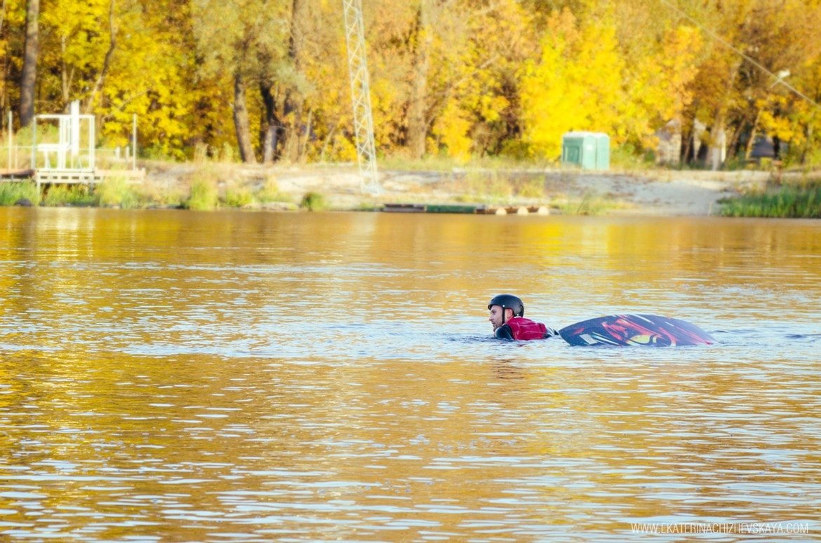 A wakeboarder wearing a black helmet and life vest floating in calm lake water with a vivid yellow autumn forest in the background.
