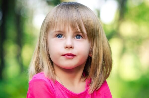Close-up portrait of a young girl with blonde hair, bangs, and blue eyes wearing a bright pink shirt outdoors with a blurred green background.