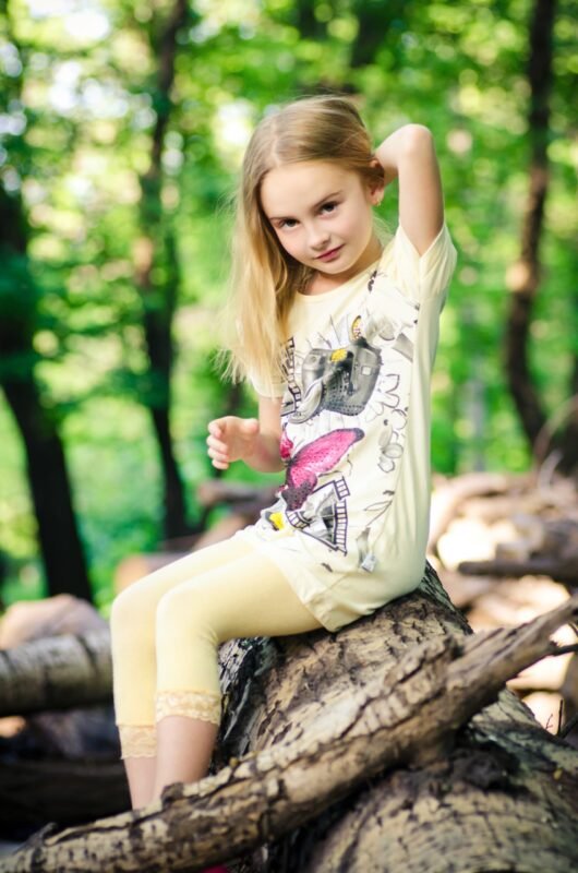 Young girl with long blonde hair sitting on a fallen log in a green forest, wearing a yellow t-shirt and matching leggings.