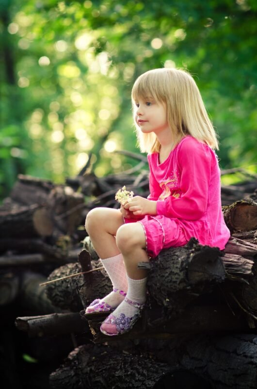 A little girl with bangs wearing a bright pink dress sitting on a pile of logs in a green park