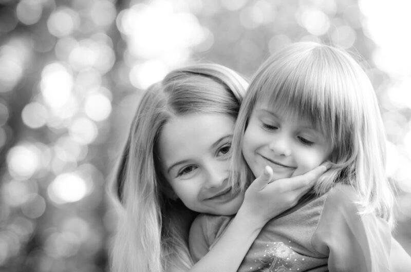 Black and white close up portrait of two young sisters hugging and smiling with a blurred background