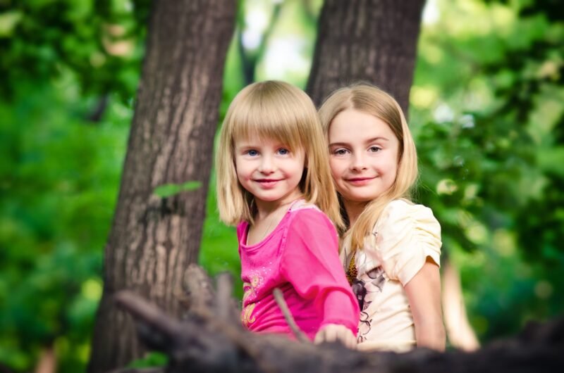 Two smiling young sisters posing together behind a fallen tree in a green summer forest