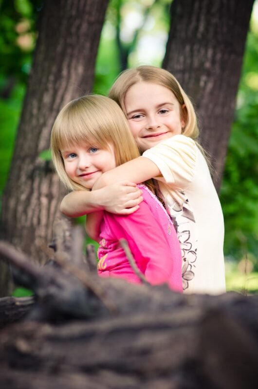 An older blonde girl hugging her younger sister from behind while posing outdoors in a forest