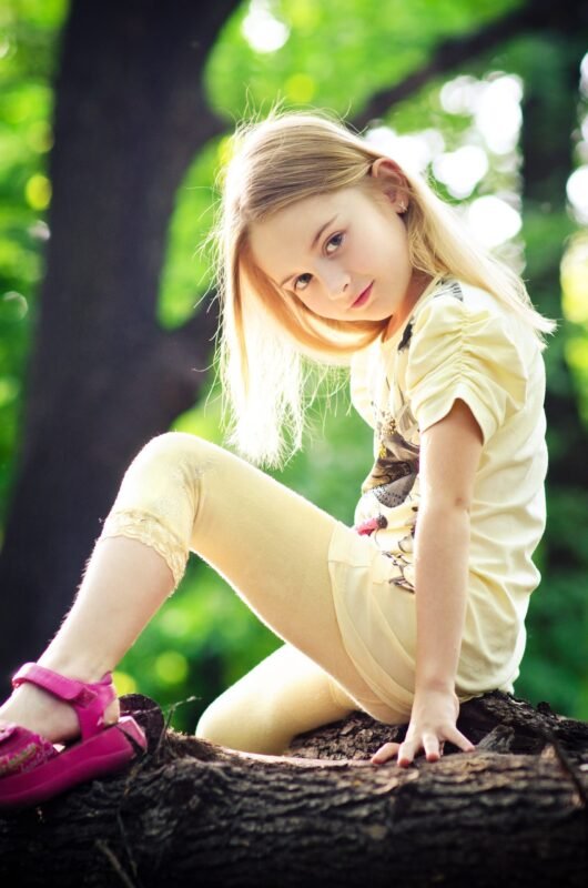 A blonde girl in a yellow outfit and pink sandals sitting on a fallen tree trunk in a sunlit park