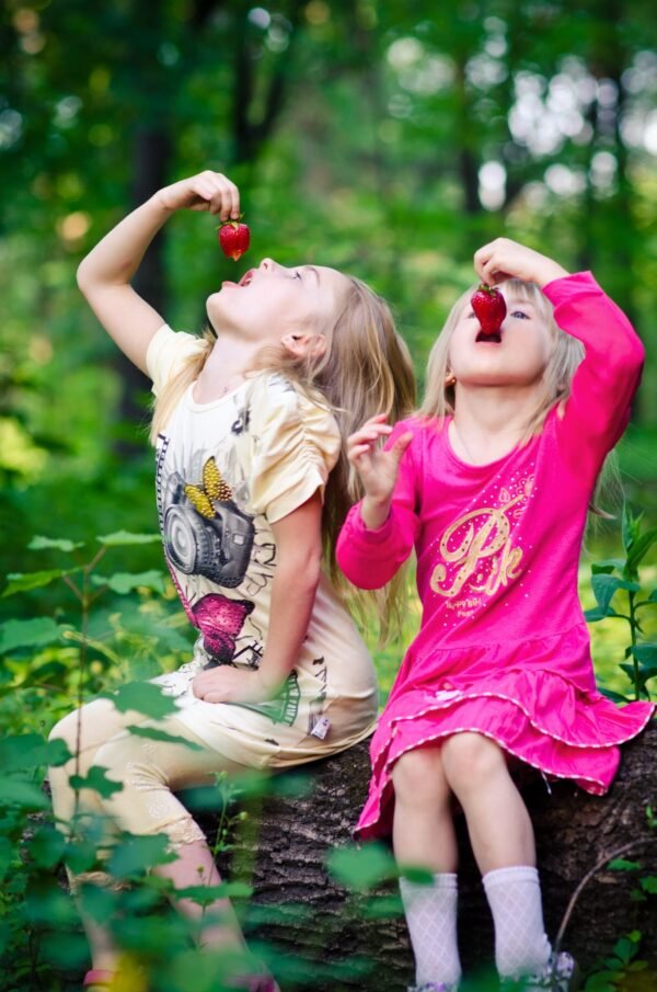 Two young girls sitting on a log in the woods and eating fresh strawberries together