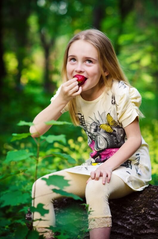 A young blonde girl in a light yellow t-shirt sitting on a log in the forest and eating a fresh strawberry