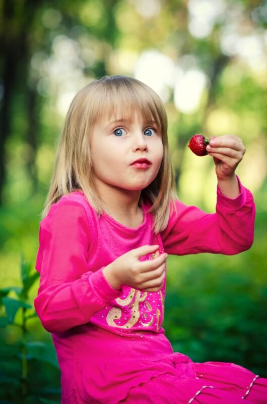A little girl in a pink dress looking surprised while holding a ripe strawberry in her hand