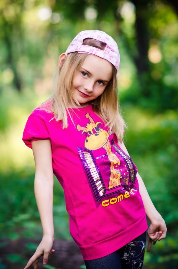 A smiling blonde girl posing in a bright pink giraffe t-shirt and a pink baseball cap outdoors