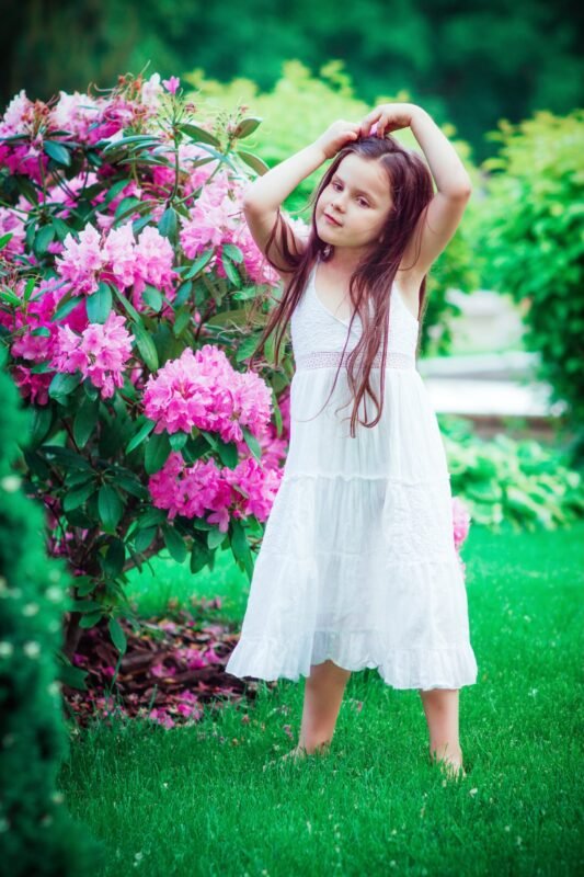 A young girl in a white summer dress posing on green grass next to a blooming pink rhododendron bush