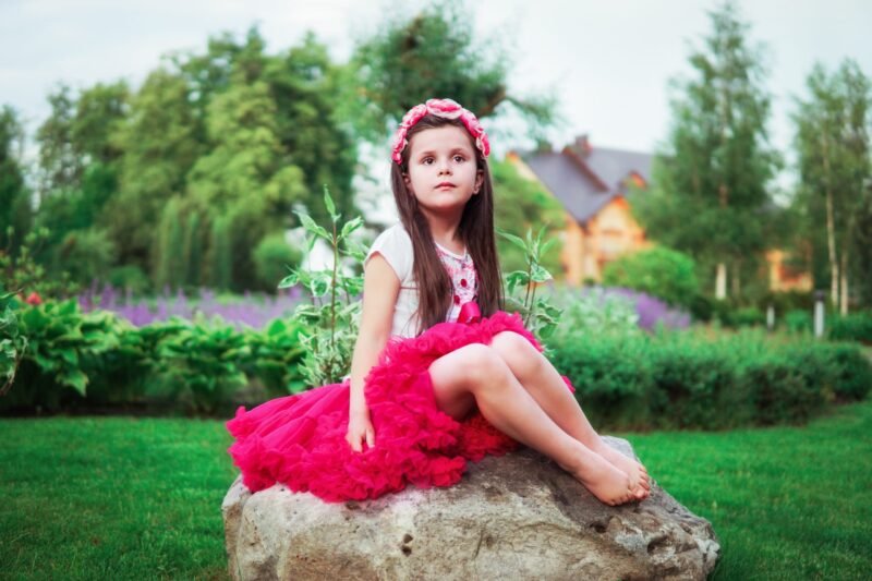 A little girl in a fluffy pink skirt and flower headband sitting on a large rock in a beautiful garden