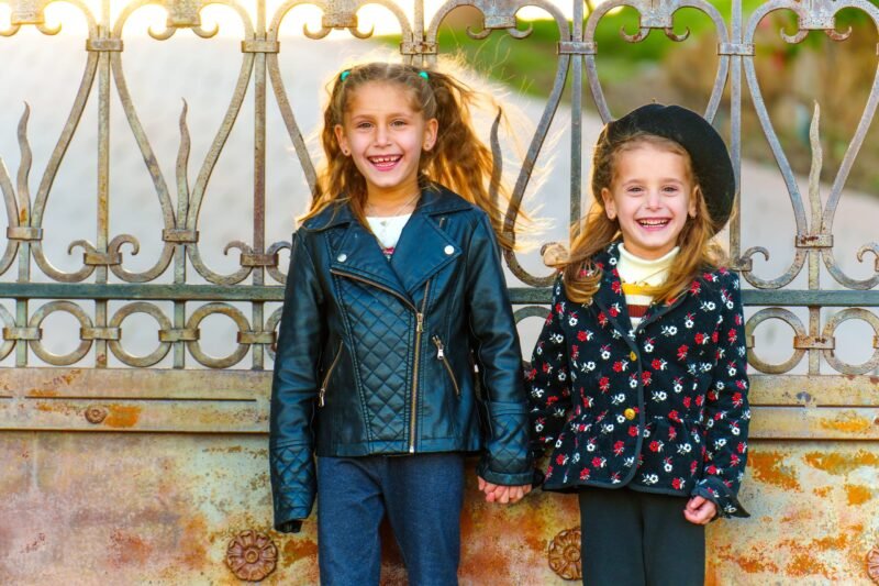 Two smiling young girls standing hand in hand next to a decorative metal fence, one wearing a leather jacket and the other a floral coat with a beret