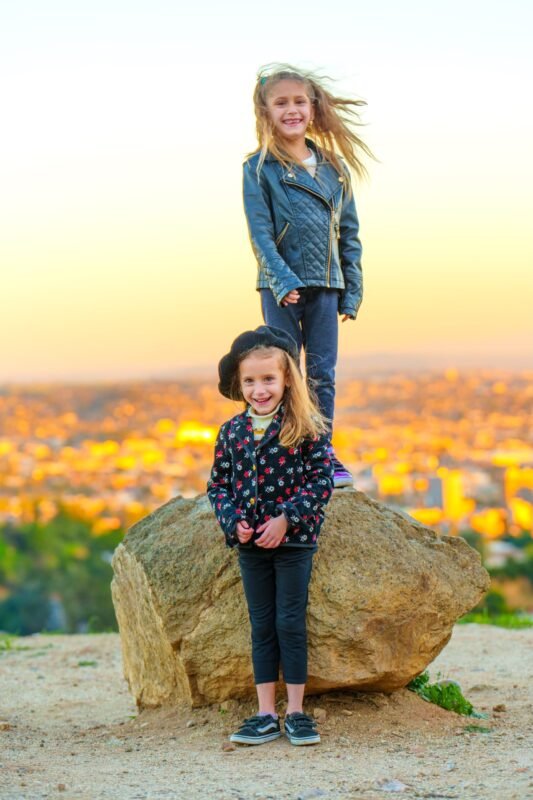 Two happy young girls posing on a large rock at sunset with a blurry city panoramic view in the background