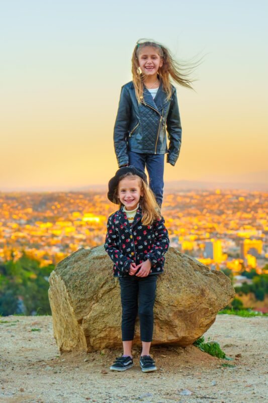 Two smiling sisters posing on a boulder overlooking a city skyline at sunset with wind-blown hair