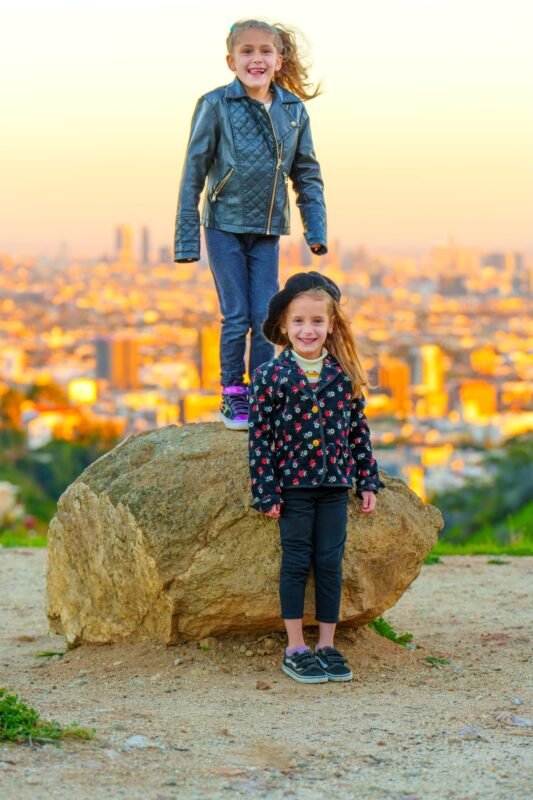 Two cheerful young girls posing by a large stone at a scenic city viewpoint during golden hour