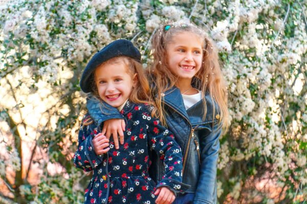 Two young girls smiling and hugging in front of a blooming white flower bush, one wearing a floral coat with a black beret and the other in a black leather jacket.