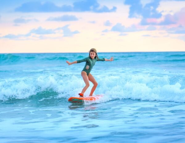 A young girl surfing on a small ocean wave during sunset, wearing a green long-sleeve swimsuit and standing balanced on an orange surfboard.