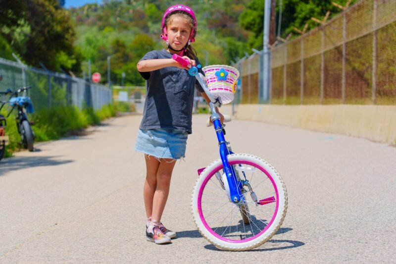 A young girl standing next to her blue bicycle with a colorful basket, wearing a pink helmet, grey shirt, and denim skirt on a sunny day.