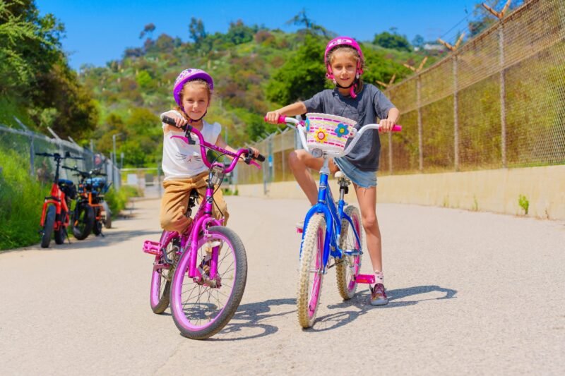 Two young girls wearing helmets posing with their bicycles on a sunny paved path with green hills and a fence in the background.