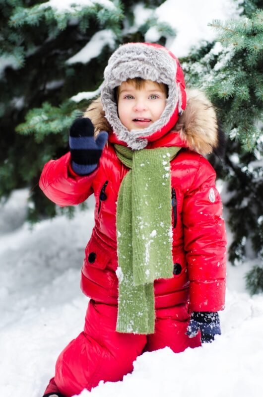 A child wearing a red winter jumpsuit, furry trapper hat, and green scarf waving a gloved hand while standing in the snow near pine trees.
