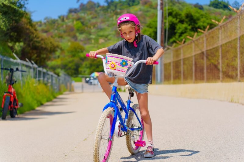 A young girl in a pink helmet and grey t-shirt balancing on a blue bicycle on an outdoor paved road.