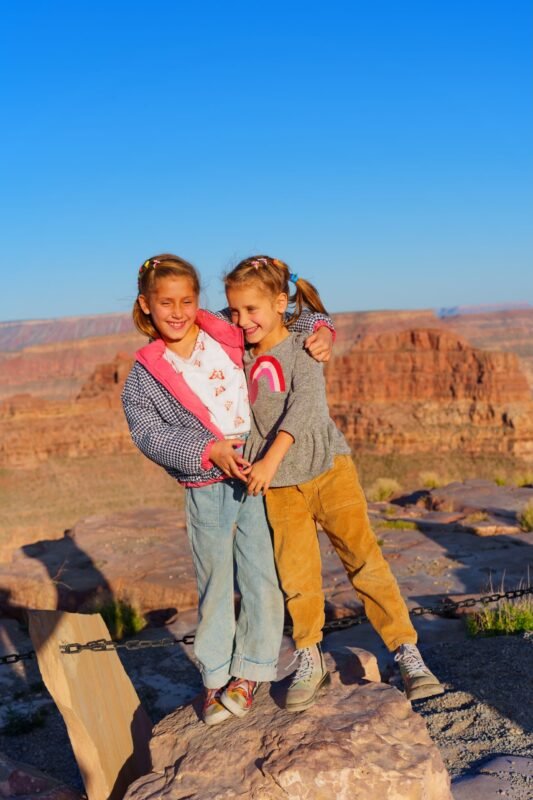 Two young girls laughing and hugging while standing on a rock at a Grand Canyon viewpoint under a clear blue sky.