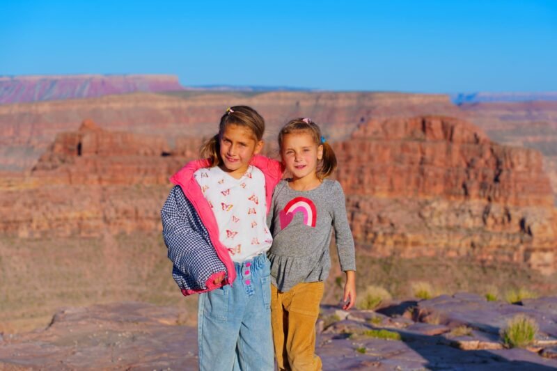 Two young sisters posing together on a rocky cliff edge with the scenic red rock canyon landscape in the background.