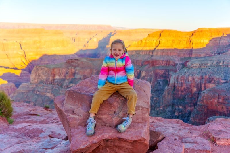 A young girl smiling while sitting on a large rock at a canyon overlook, wearing a bright rainbow-colored puffer jacket.