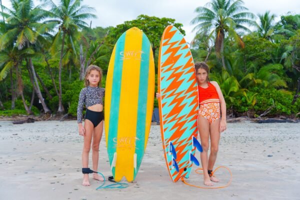 Two young girls holding surfboards while standing on a sandy tropical beach with palm trees in the background.