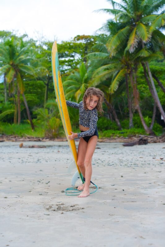 A young girl with wet hair standing on a sandy beach holding a long yellow and green surfboard near palm trees.