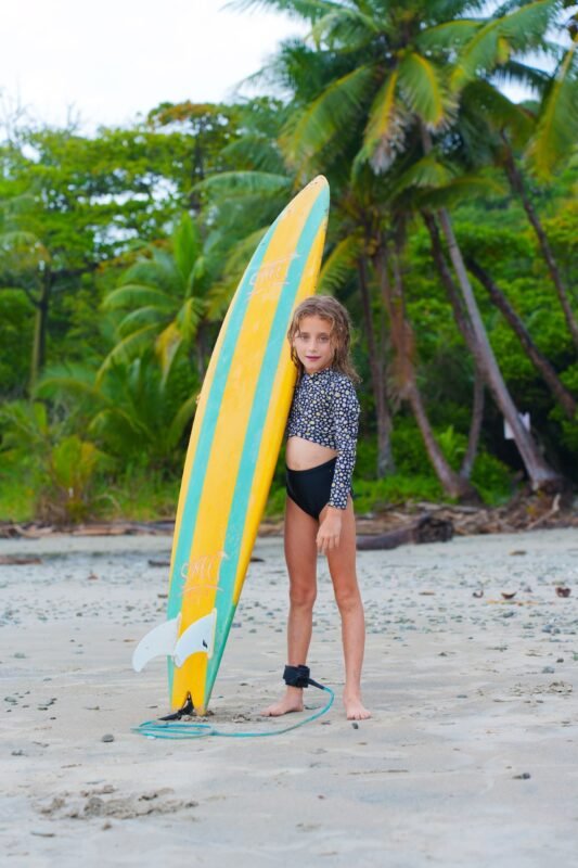 A young girl posing on a tropical sandy beach next to a tall yellow surfboard with a leash tied to her ankle.