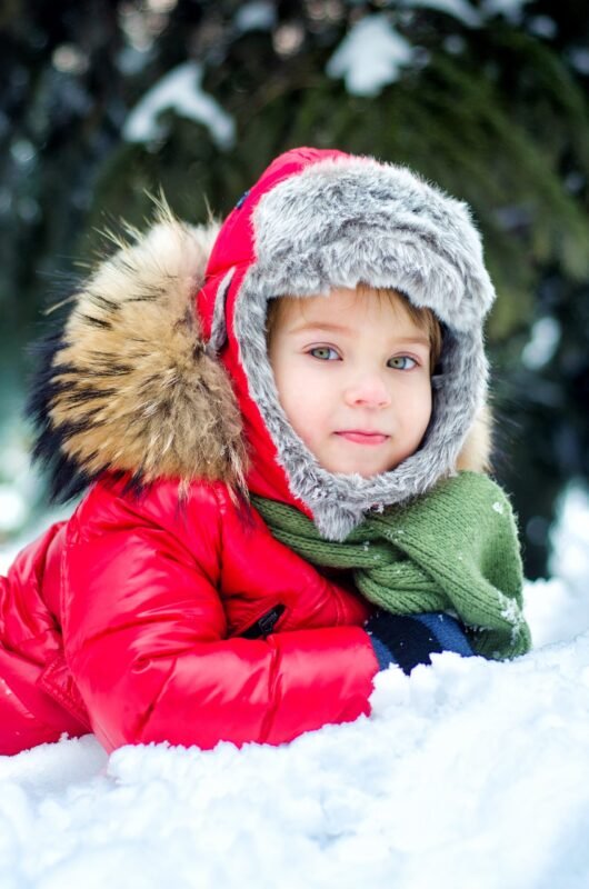Close-up of a young child lying in the snow wearing a bright red winter coat with a fluffy hood and a green scarf.
