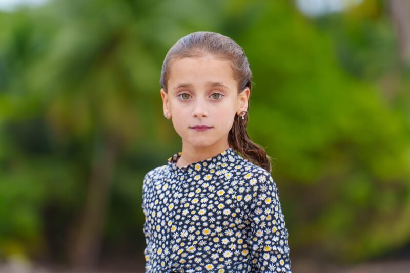 Close-up portrait of a young girl with brown eyes wearing a floral long-sleeve shirt against a blurred green background.