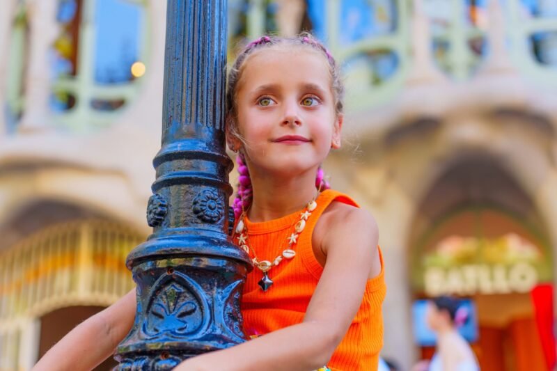 A young girl with braided hair and a seashell necklace leaning against a black lamppost outdoors.