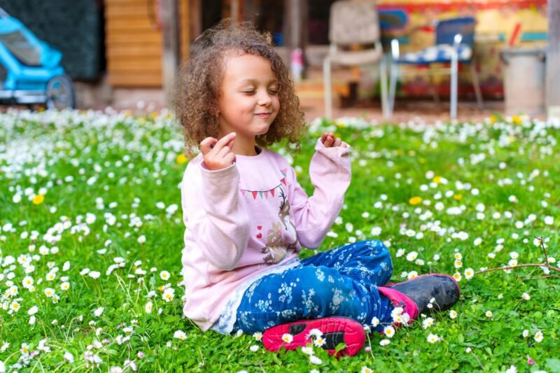 A happy young girl with curly hair sitting cross-legged on a green grass field full of small white daisy flowers.