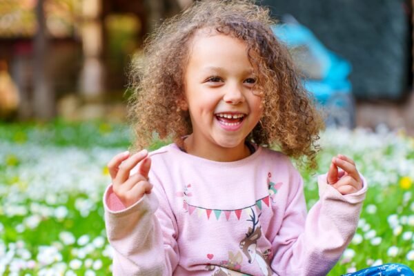 A smiling young girl with curly hair wearing a pink sweater, sitting in a flower meadow and gesturing with her hands.
