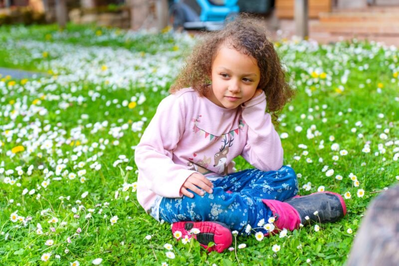 A young girl with curly hair sitting on the grass thoughtfully with her hand on her cheek in a field of daisies.