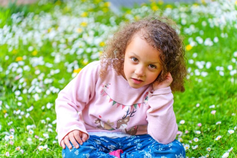 A young girl with curly hair looking directly at the camera while sitting in a green field with white wildflowers.