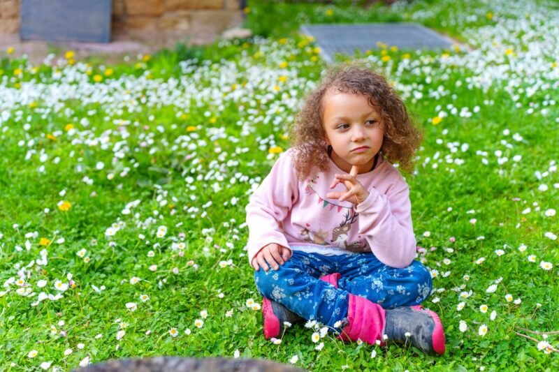 A young curly-haired girl in a pink sweatshirt and blue floral pants sitting cross-legged on a green lawn covered with white daisies.