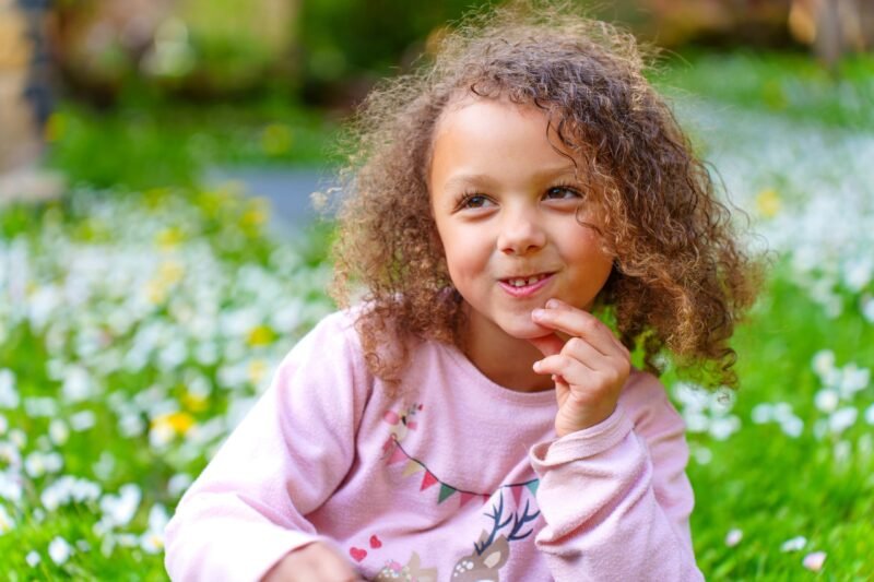 Close-up portrait of a cheerful young girl with curly hair sitting in a green grassy field filled with white wildflowers, looking away thoughtfully.