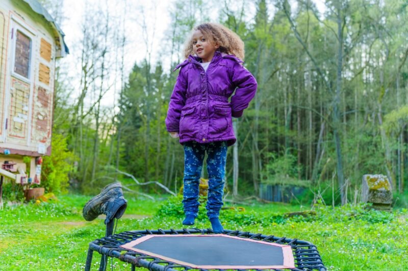 A young girl with curly hair in a purple winter jacket jumping high on a small outdoor fitness trampoline in a green backyard near a forest.