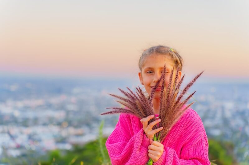 A young girl with braided hair wearing a bright pink sweater and holding a bunch of decorative grass field stalks with a blurred city landscape background at sunset.