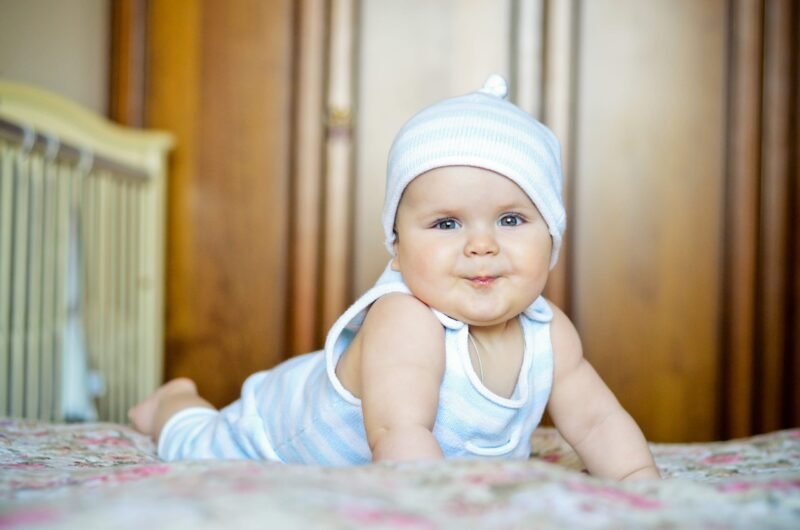 Cute baby wearing a blue and white striped hat and matching sleeveless outfit lying on a floral blanket while smiling.